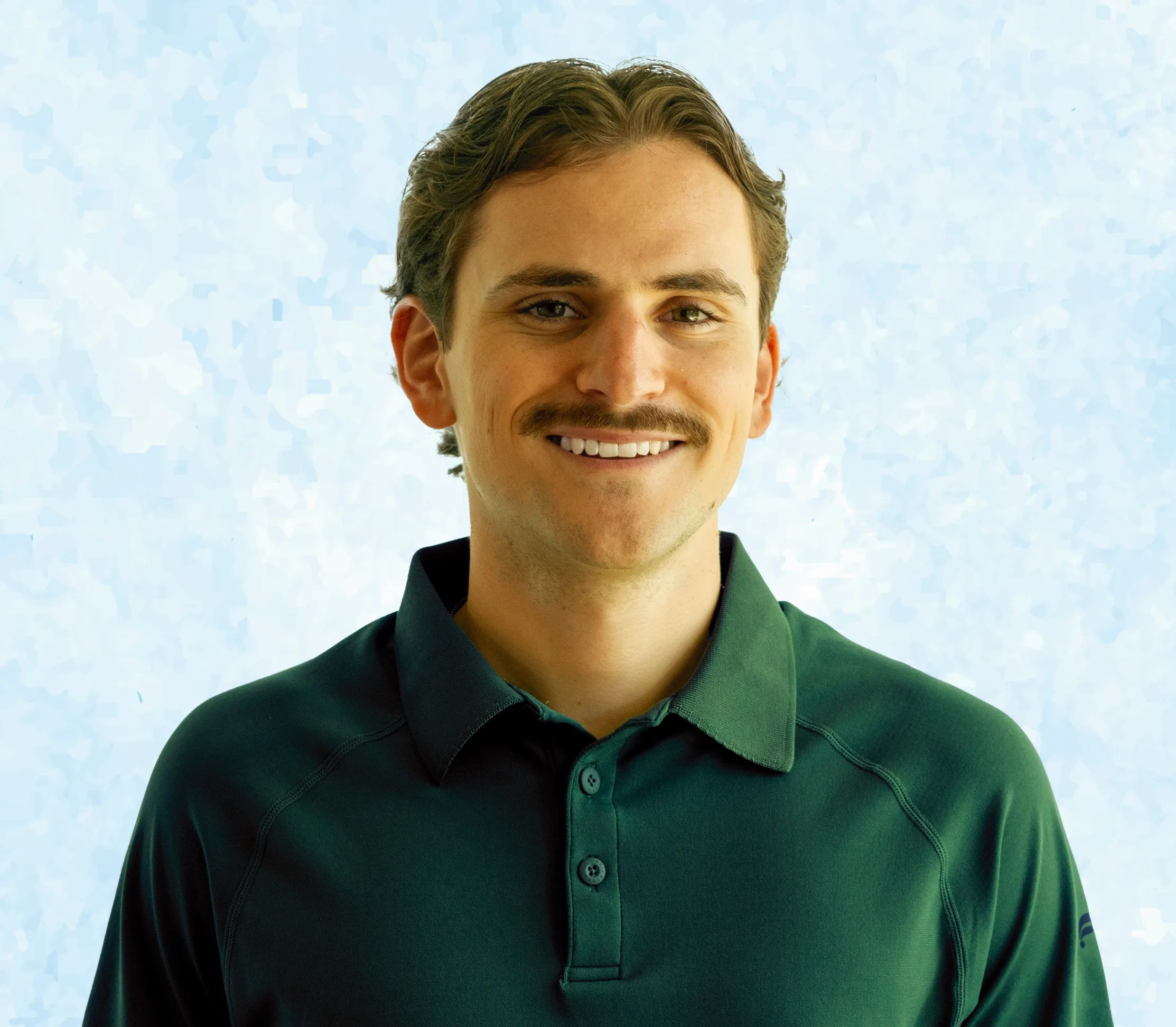 Smiling man with mustache wearing green collared shirt against light blue background.